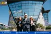 Three individuals in business attire and hard hats stand confidently in front of a modern glass building. The individuals appear to be construction professionals, with one holding a blueprint and another wearing a safety vest. The background features a clear blue sky and landscaped greenery.