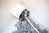 Dryer vent in a home being cleaned out with a round brush. There is a large pile of lint that has been removed from the vent on a white tiled floor. The walls and baseboards are white. The lint is gray. 
